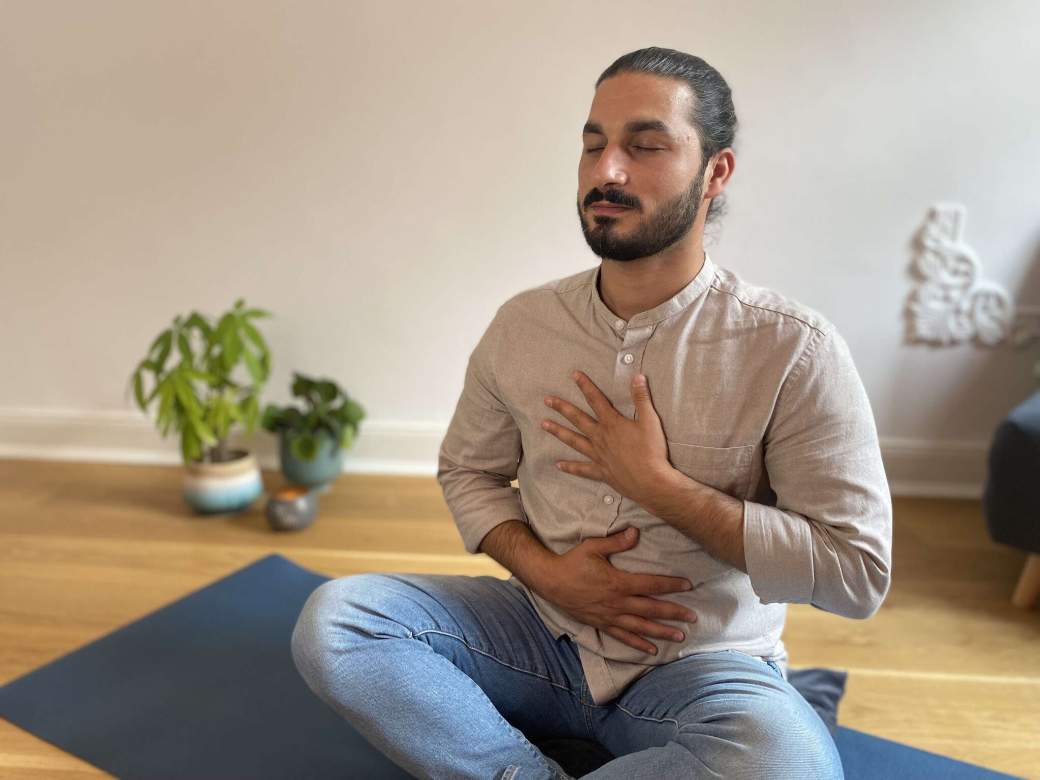 Person sitzt meditierend auf einer Yogamatte in einem Raum mit Pflanze im Hintergrund.